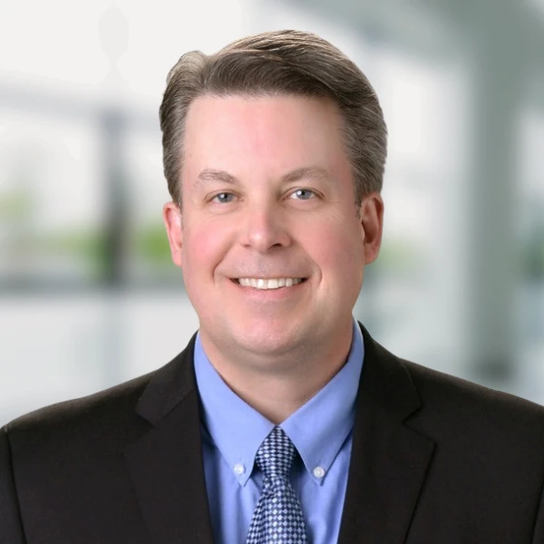 Man smiles in front of a blurred background. He is wearing a suit and tie.