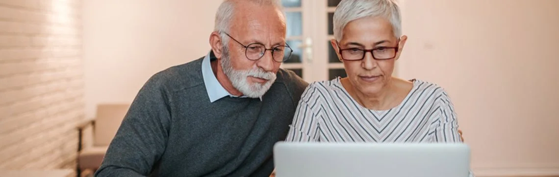 Elderly couple focused on a laptop screen, sitting close together. The man wears glasses and a sweater, the woman wears glasses and a striped blouse.