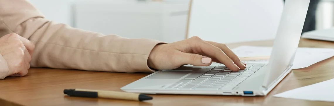 A person in a beige jacket types on a laptop at a wooden desk, with a pen and papers nearby, conveying a productive, focused atmosphere.