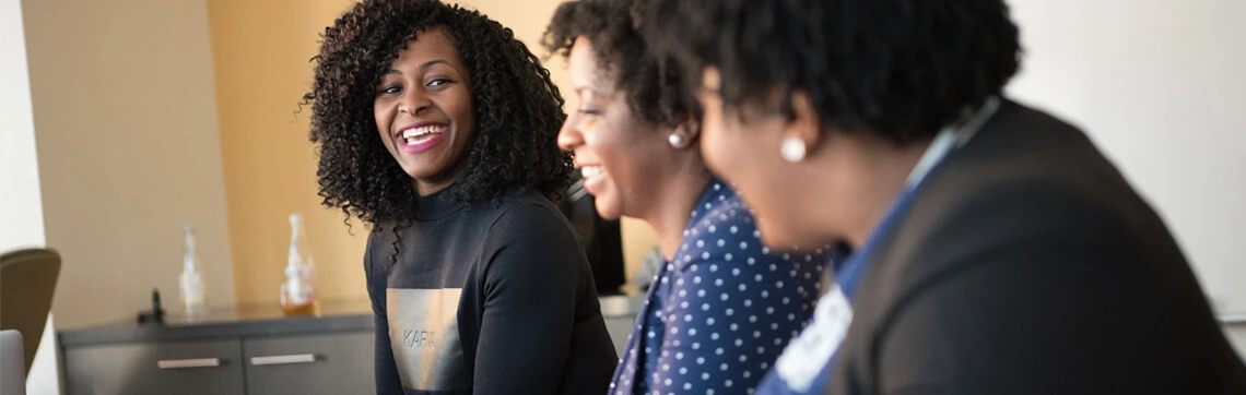Three women sitting together and smiling during a casual conversation in an office setting.
