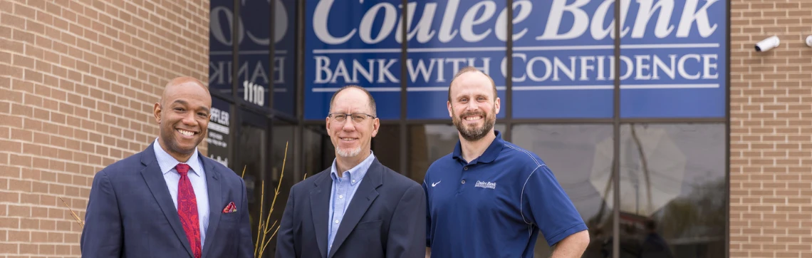 Three men stand smiling in front of a bank. The first wears a suit with a red tie, the second a suit jacket and glasses, the third a blue polo.