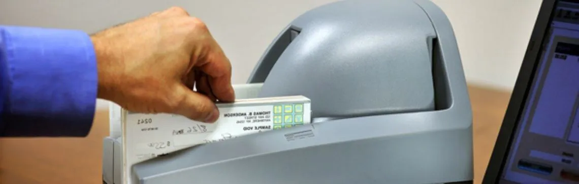 Hand inserting checks into a desktop check scanner next to a computer for remote deposit.