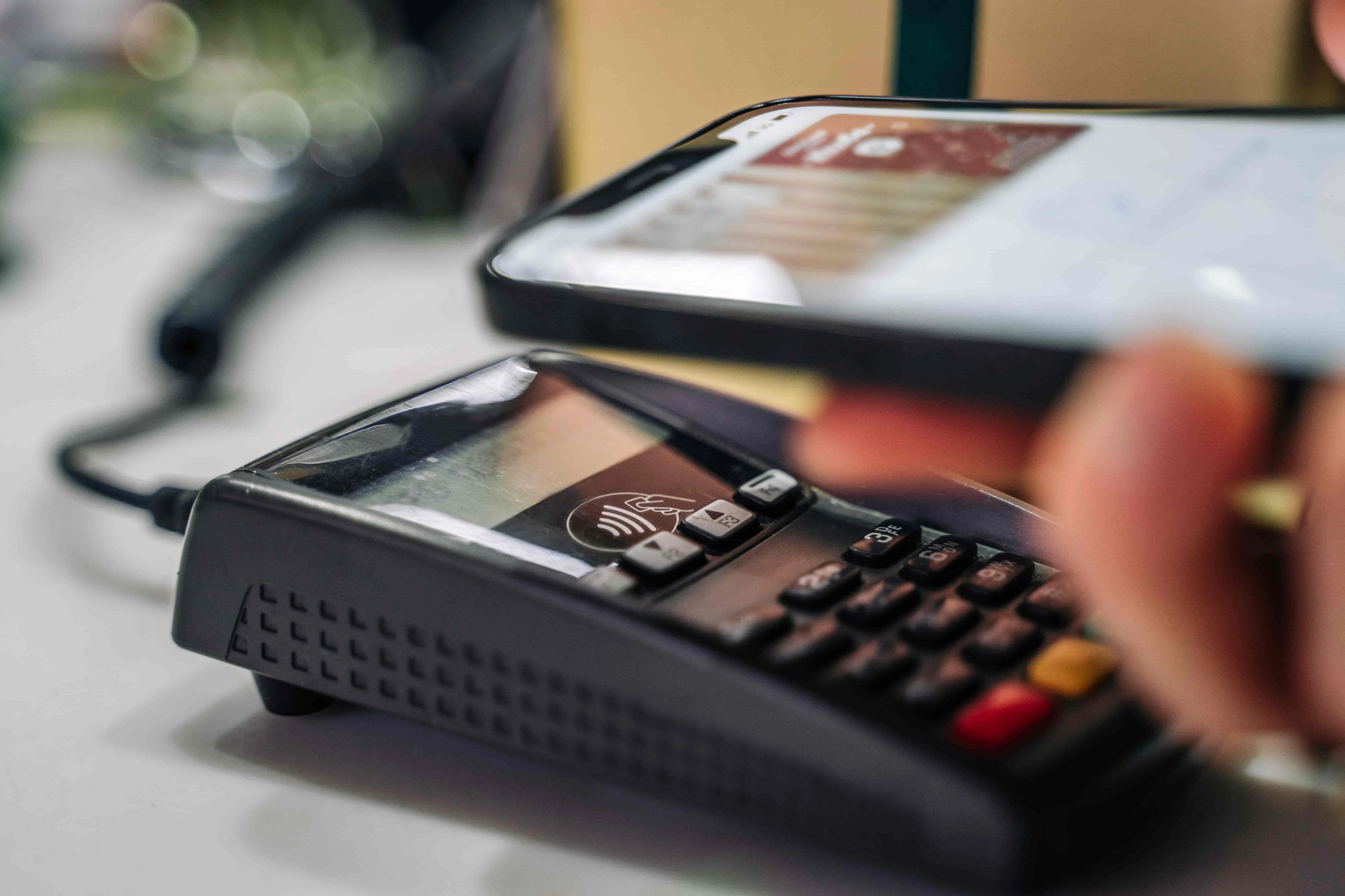 A hand holds a smartphone above a card payment terminal, illustrating a contactless payment in a retail setting. The focus is on technology and convenience.