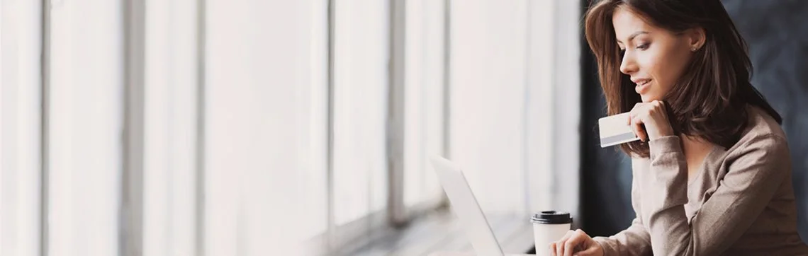 A woman seated at a table looks at a laptop screen, holding a credit card. She appears focused and relaxed. A coffee cup is beside her, with soft window light streaming in.