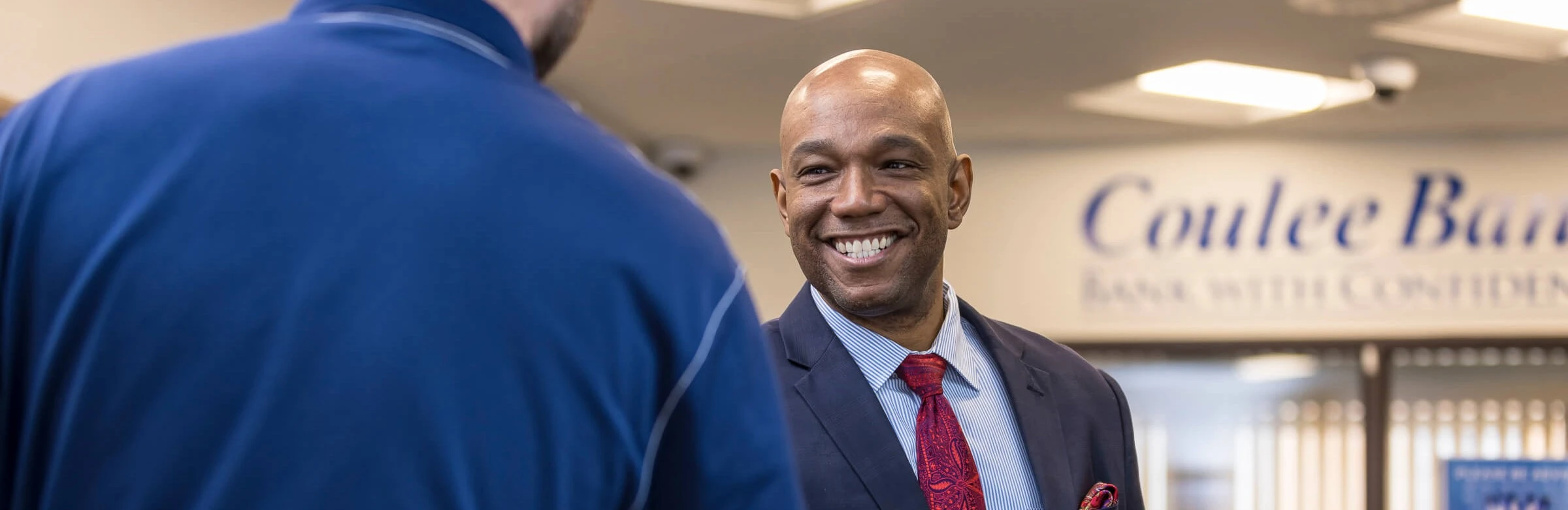 Smiling man in a suit talking with another person inside a bank.